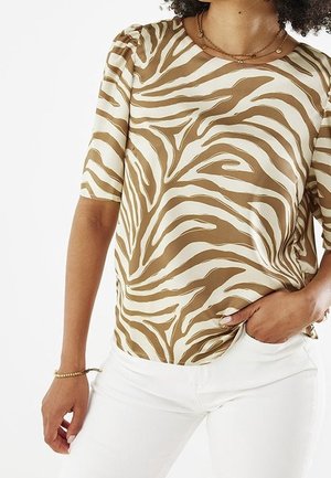 Woman wearing a tan and white zebra print short-sleeve blouse with puffed shoulders and white pants, hand in pocket, showing gold jewelry.