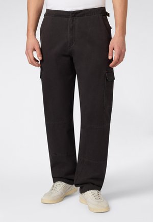 Homme portant un pantalon cargo noir ample avec des poches latérales et des baskets blanches, debout devant un fond clair uni.