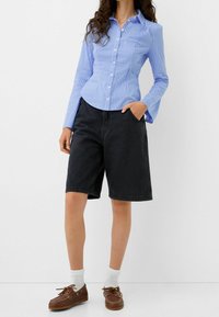 Jasnoniebieska koszula w paski z długim rękawem, zestawiona z czarnymi szortami do kolan i brązowymi butami typu boat shoes w białych skarpetkach.