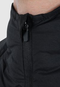 Veste noire avec un col montant et une fermeture éclair visible. Fabriquée en tissu lisse et léger avec une texture mate. Tirette de la fermeture éclair en noir.
