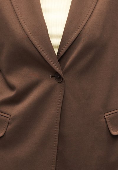 Κοντινή λήψη ενός καφέ σακακιού με ένα κουμπί, που δείχνει ραμμένους γιακάδες, εμπρόσθιες τσέπες με καπάκια, πάνω από ένα ανοιχτό μπεζ ριμπ πλεκτό τοπ.