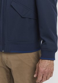 Navy zip-up jacket with textured fabric, featuring a slanted pocket and contrasting blue trim at the hem and cuffs. Hands visible at the side.