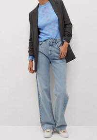 Grijze blazer over een lichtblauwe, met patroon gebreide trui, gecombineerd met licht gewassen wijde jeans en witte schoenen met crèmekleurige accenten.