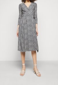 Schwarz-weißer gemusterter Kleid mit langen Ärmeln, V-Ausschnitt und ausgestellt Rock. Getragen mit hellen, strapazierfähigen Sandalen.