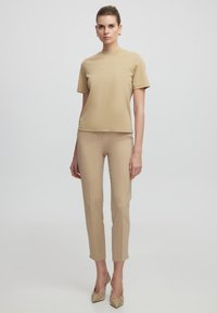 Beige krátký rukáv top a dobře padnoucí kalhoty ve stejném odstínu, vyrobené z hladkého materiálu. Modelka má na sobě texturované béžové lodičky. Jednoduchý, moderní design.