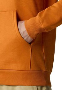 Sudadera naranja con bolsillo canguro, puños y dobladillo acanalados. La tela es suave y texturizada, con un diseño de ajuste relajado.