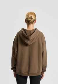 Felpa marrone oversize con una texture morbida e un grande cappuccio, caratterizzata da spalle scese e polsini a coste. Design semplice senza motivi.