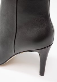 Gros plan d'une bottine noire en cuir à talon haut montrant une texture lisse, des coutures et un talon fin sur fond blanc.