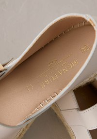 Espadrille-Schuh mit einem hellbeigen Lederoberteil, weicher Innenausstattung und Espadrille-Seilsohle; mit dezentem Branding in Goldfolie.