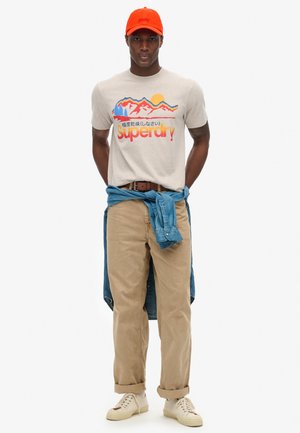 T-shirt graphique beige avec un motif de montagne, pantalon en velours côtelé marron clair, chemise en denim nouée autour de la taille, casquette orange et baskets crème.