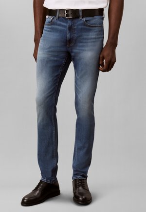 Homme portant un jean bleu slim-fit, une ceinture en cuir noire, une chemise blanche et des chaussures habillées noires, debout devant un fond uni.