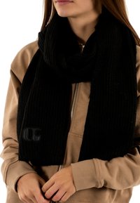 Écharpe noire en tricot avec une texture côtelée et un accent logo, drapée autour du cou ; associée à un sweat à capuche beige.