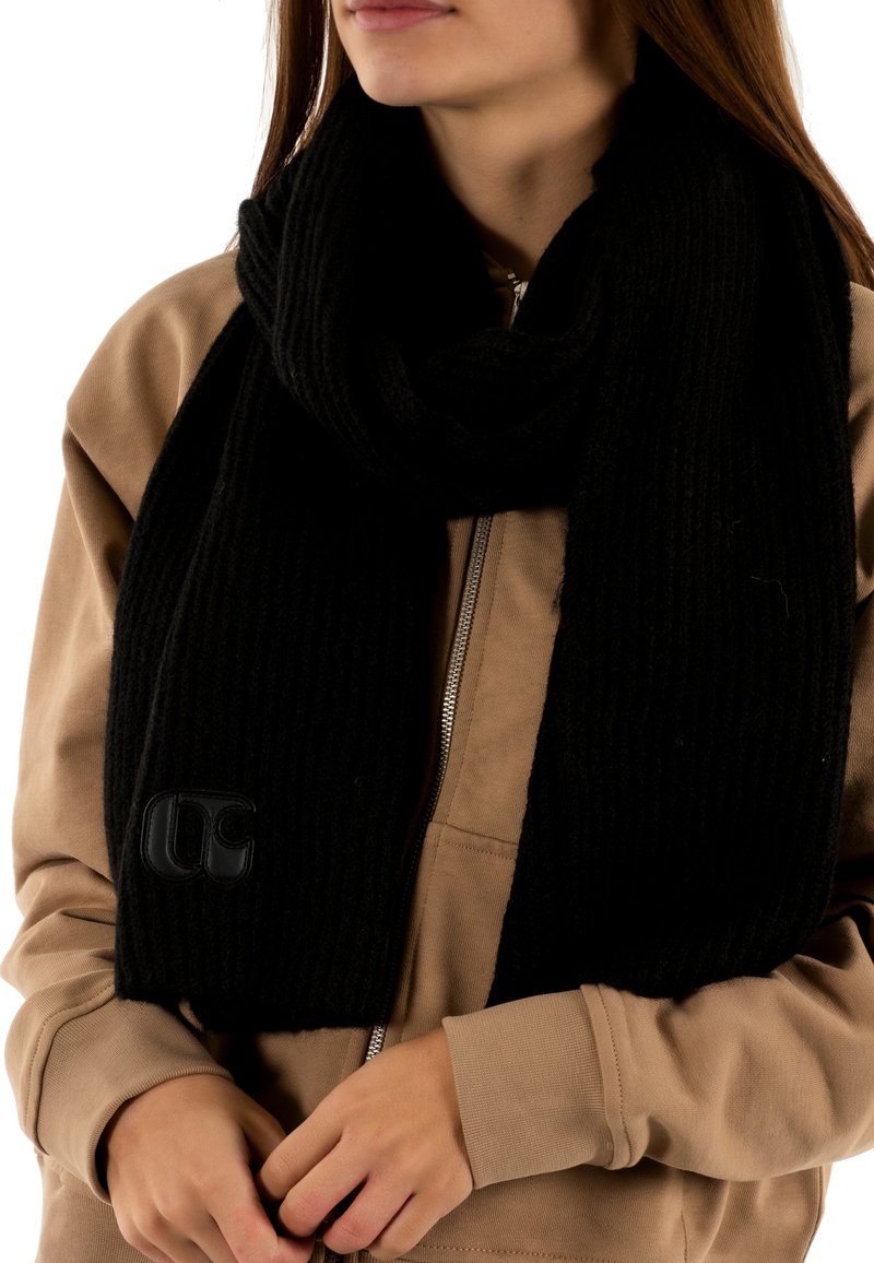 Écharpe noire en tricot avec une texture côtelée et un accent logo, drapée autour du cou ; associée à un sweat à capuche beige.