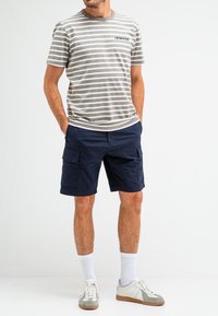 Gestreift graues und weißes T-Shirt mit Rundhalsausschnitt, kombiniert mit marineblauen Cargo-Shorts. Das Model trägt weiße Socken und grau-weiße Turnschuhe.