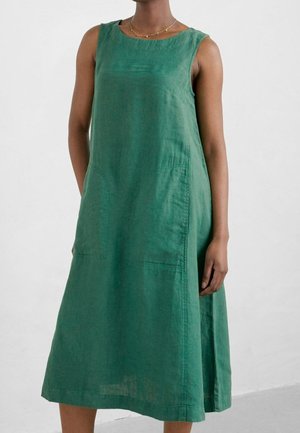 Mujer vestida con un vestido midi verde suelto sin mangas, con grandes bolsillos delanteros y un collar delicado, de pie frente a un fondo blanco liso.