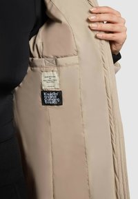 Interiér béžovej prešívanej bundy s látkovou nálepkou s čiernym textom a druhou nálepkou v čiernej a bielej farbe. Hladká textúra a prešívaný dizajn.