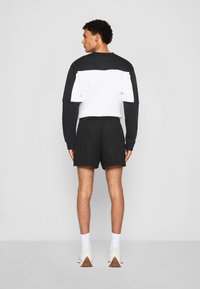 Sweatshirt noir et blanc à blocs de couleur avec des manches longues, associé à un short en mesh noir. Des chaussettes blanches et des chaussures de sport beiges complètent le look.
