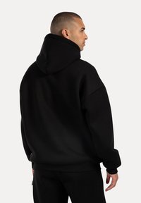 Mann mit kurzem Haar, der einen oversized schwarzen Hoodie trägt, seitlich gegen einen schlichten hellen Hintergrund blickend.