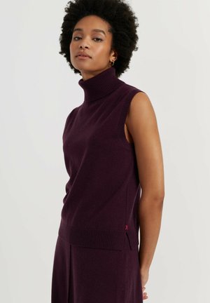 Vrouw met kort krullend haar, gekleed in een mouwloze donkerpaarse coltrui en bijpassende rok tegen een effen achtergrond.
