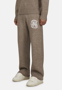 Brązowe dzianinowe spodnie dresowe o luźnym kroju, z białym logo na udzie i ściągaczami. Połączone z białymi sneakerami.