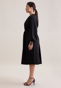 Czarna sukienka midi o kroju kopertowym, z długimi bufiastymi rękawami i paskiem w talii. Połączona z czarnymi sandałami na wysokim obcasie, gładka faktura materiału.