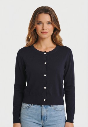 Donna con capelli castani mossi indossa un cardigan blu navy con piccoli bottoni a forma di cuore e jeans azzurri, posando contro uno sfondo bianco uniforme.