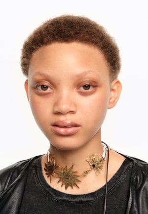 Persona joven con cabello corto y rizado que lleva una blusa negra y un collar tipo choker dorado con diseños de sol y palmeras.