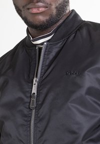 Zwarte bomberjack met ritssluiting, aansluitende kraag en geborduurd merkembleem. Gladde textuur met subtiele glans.