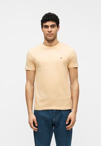 Άνδρας με σγουρά μαλλιά όρθιος, φορώντας ανοιχτό μπεζ κοντομάνικη μπλούζα και μπλε τζιν, με φόντο απλό λευκό.