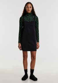 Zwarte overall jurk met een vierkante halslijn en een zak, gedragen over een groene blouse met lange mouwen en bloemenprint. Zwarte sokken en schoenen maken de outfit compleet.