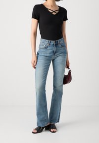 Femme portant un top noir à encolure croisée, jean taille haute bleu clair, sandales noires ouvertes aux orteils, tenant un petit sac à main foncé.