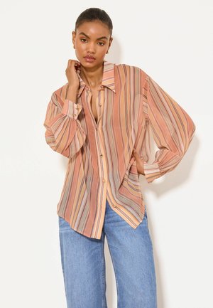 Žena nosi voľnú broskyňovo a béžovo pruhovanú košeľu s širokými rukávmi a modré džínsy, pózuje s rukou blízko tváre a bokov.