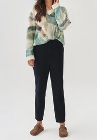 Sweter z dzianiny z dekoltem w serek w kolorze morskim i kremowym, w połączeniu z ciemnogranatowymi spodniami. Model nosi brązowe buty wsuwane. Miękka faktura i luźny krój.