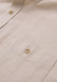Beige-linen-paita, jossa on nappi rintataskussa, tekstuurinen kangas ja ompeluyksityiskohtia taskun reunoilla.