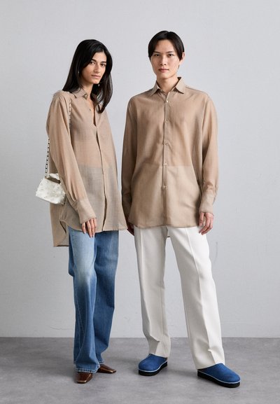 Due persone indossano camicie button-up di un colore marrone chiaro in un tessuto morbido. Una persona ha dei jeans blu sbiaditi, l'altra indossa pantaloni bianchi e scarpe blu.