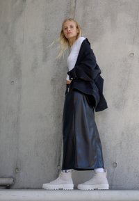 Zwarte leren maxi rok, witte lange mouw shirt en crèmekleurige high-top sneakers. Model poseert tegen een betonnen muur.