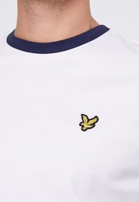 Vit bomull t-shirt med en mörkblå ribbad rundhals, med en liten gul broderad fågel-logotyp på bröstet.