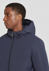 Giacca con cappuccio blu navy con zip, realizzata in un tessuto liscio e testurizzato. Caratterizzata da un patch con logo sulla spalla.