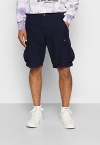 Homme portant un short cargo bleu marine, des baskets montantes blanches et un sweat-shirt à manches longues tie-dye avec du texte sur un fond blanc.