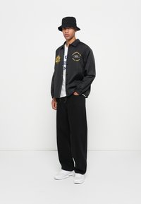 Black lightweight jacket with gold lettering, paired with a gray graphic t-shirt, black wide-leg pants, and white sneakers, all set against a plain background.