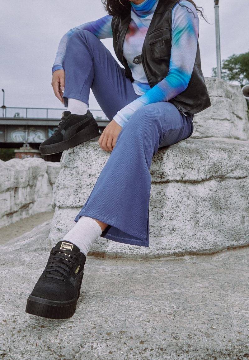 Černé semišové platformové tenisky s tlustou podrážkou. Oblečení zahrnuje dlouhý rukáv s modrým tie-dye vzorem, černou koženou vestu a modré zvonové kalhoty.