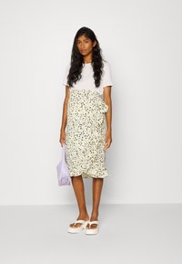 Top branco de manga curta emparelhado com uma saia amarela com padrão floral, apresentando uma bainha em babados e um nó lateral. Inclui também uma bolsa roxa e sandálias de plataforma brancas.