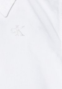 Balta medvilnės marškiniai su audiniu tekstūra, turi mažą išsiuvinėtą "cK" logotipą derančia spalva šalia apykaklės.