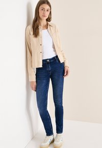 Chaqueta de pana beige sobre una camiseta blanca, combinada con jeans de mezclilla azul y zapatillas de color claro. El fondo es neutro.