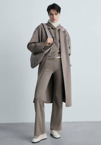 Béžový vlnený kabát cez béžovú mikinu a nohavice, skombinovaný so svetlými topánkami. Veľká, textúrovaná taška na rameno dotvára outfit.