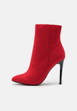 Bottine en daim rouge avec bout pointu et talon stiletto noir sur un fond clair uni.