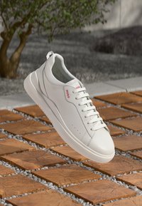 Białe skórzane sneakersy z okrągłym noskiem, płaskimi sznurowadłami i czerwonym logo "HUGO". Gumowa podeszwa z teksturowanym wzorem, położona na drewnianych płytkach.