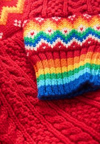 Červený pletený svetr s texturovaným vzorem a barevnými žebrovanými manžetami v duhových odstínech, včetně modré, zelené a oranžové.