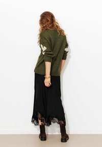 Pull en maille vert olive avec des accents blancs, associé à une jupe noire avec des bordures en dentelle. Des bottines marron à la cheville complètent l'ensemble.