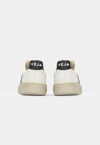 White leather sneakers with black accents, featuring a textured heel, beige rubber sole, and "VEJA" logo on the back.